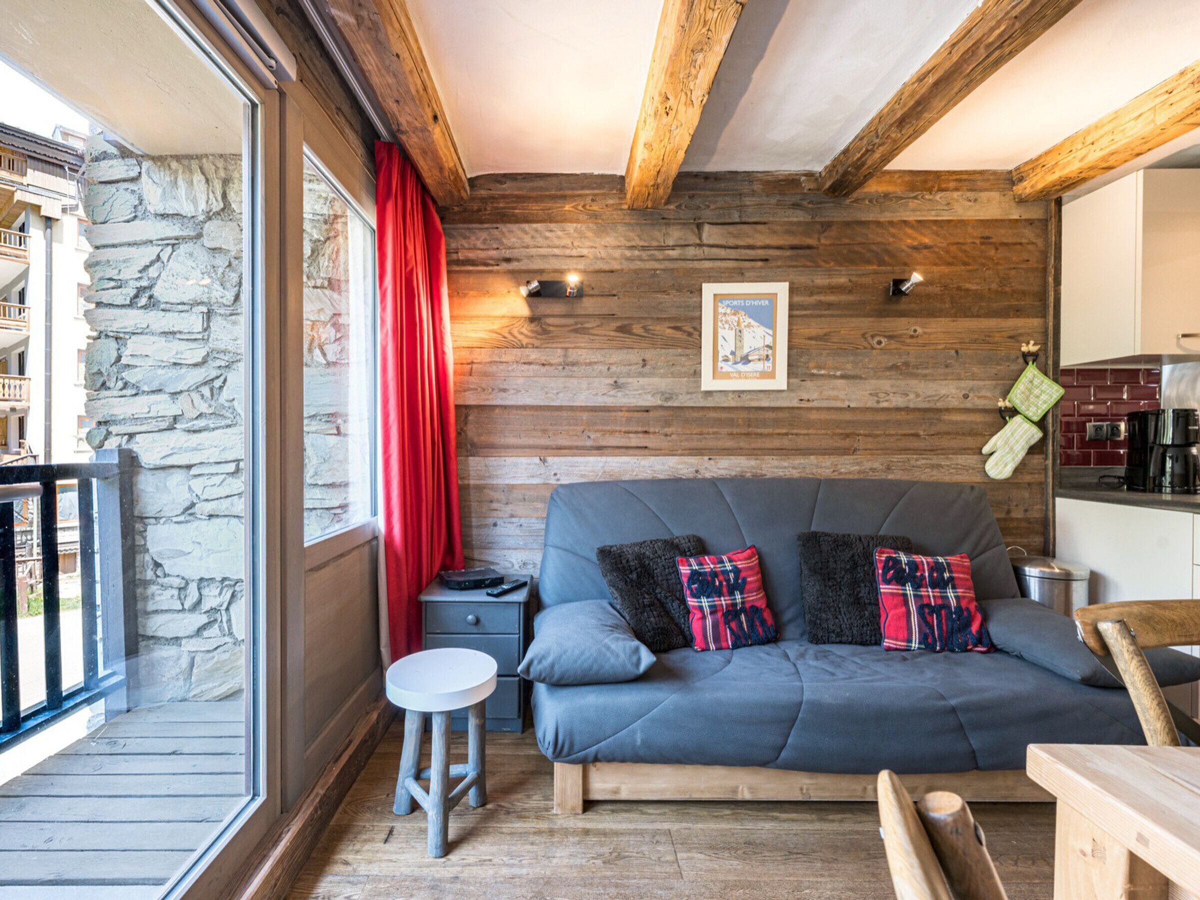 A cozy room with a stone wall, wooden ceiling beams, a grey sofa with plaid cushions, a small round table, and a view through a glass door.
