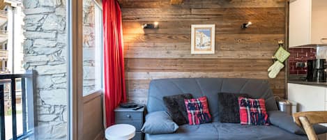 A cozy room with a stone wall, wooden ceiling beams, a grey sofa with plaid cushions, a small round table, and a view through a glass door.