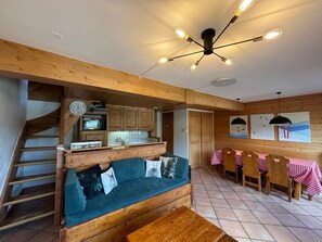 Ein gemütliches Interieur mit einer Holztreppe, einer Couch mit Kissen, einer Essecke mit Tisch und Stühlen sowie einer Küche mit Schränken und einer Mikrowelle.