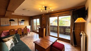 Een gezellige kamer met een houten plafond, een eettafel met stoelen, een bank, een kleine tafel en uitzicht op een houten terras buiten.