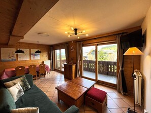 Ein gemütliches Zimmer mit Holzbalkendecke, einem Esstisch mit Stühlen, einem Sofa, einem kleinen Tisch und Blick auf eine Holzterrasse.