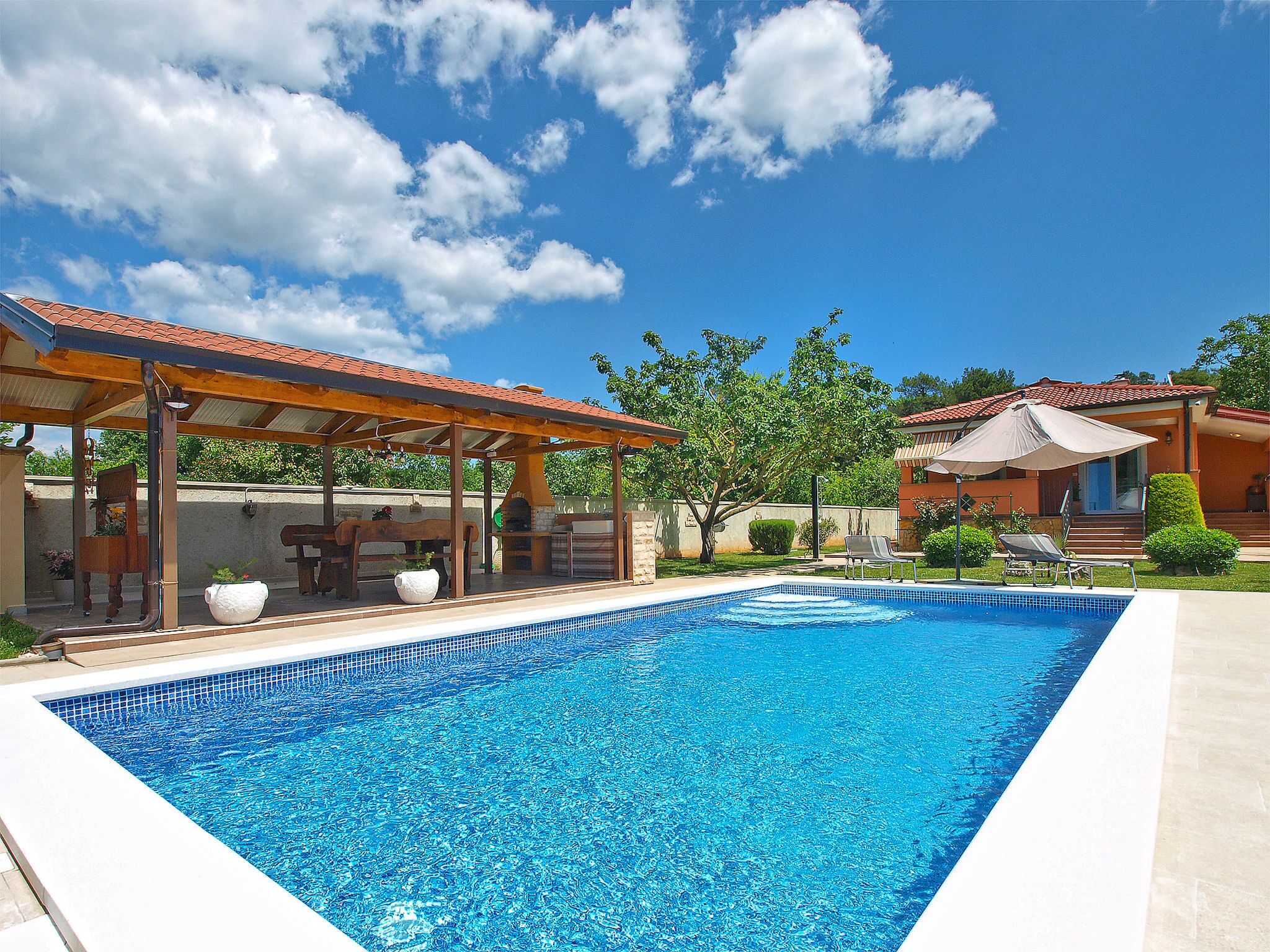 A swimming pool with a covered patio area, a clear blue sky, and surrounding greenery.