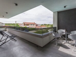 Un balcón moderno con barandilla de vidrio, sillas blancas, una mesita y una botella de vino.