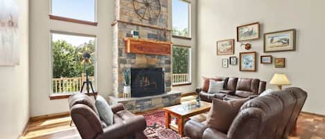 A spacious living room with a stone fireplace, leather sofas, and a large window overlooking greenery.