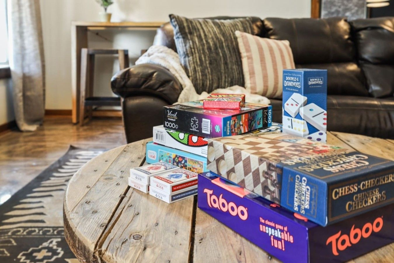 A variety of board games on a wooden coffee table in a living room with a black leather sofa and a patterned rug.