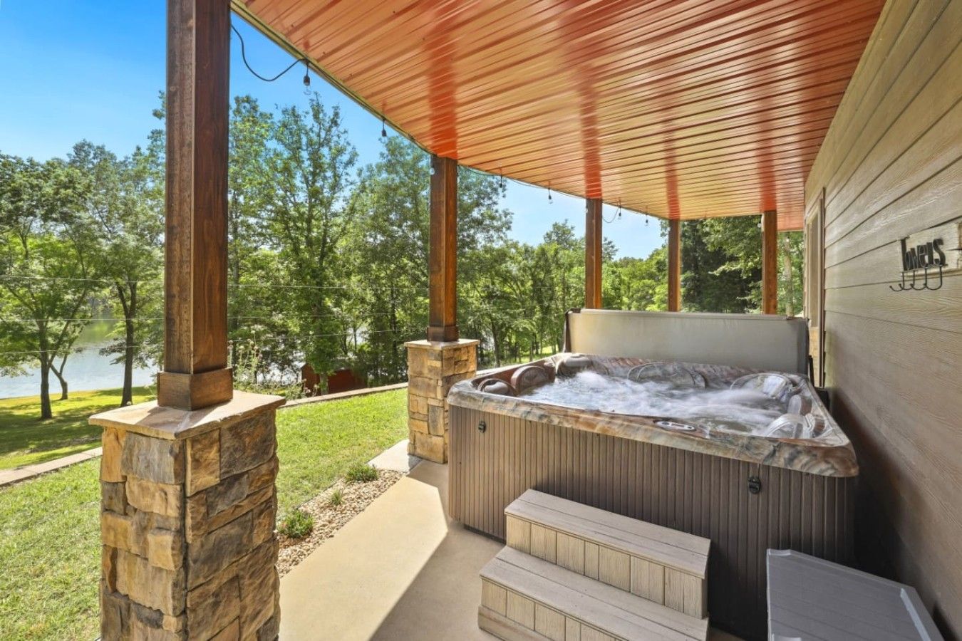 A hot tub under a wooden pergola with stone pillars, surrounded by a grassy area and trees.