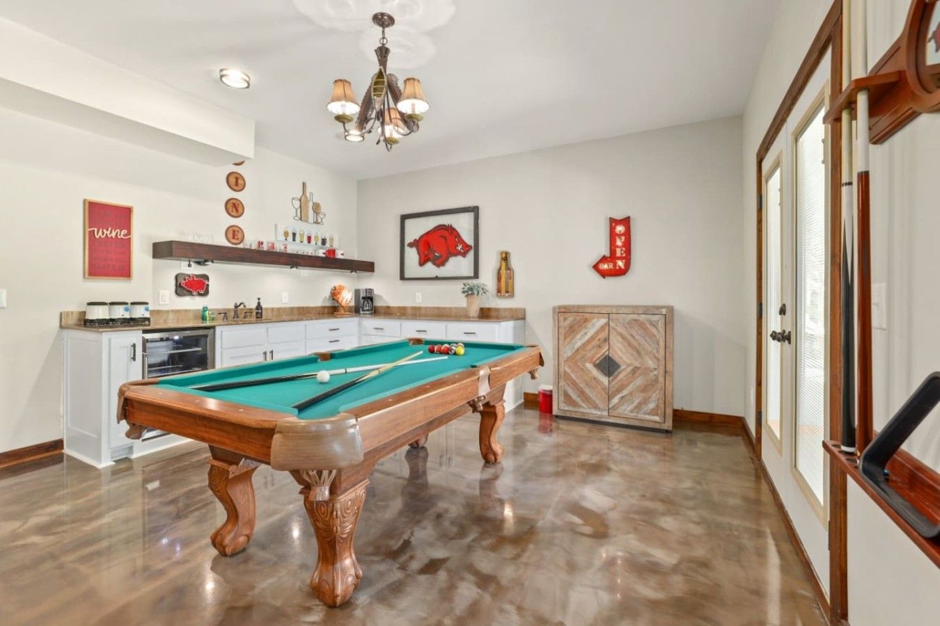 A pool table in a room with a kitchen area, a chandelier, and a door with a decorative panel.
