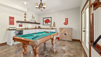 A pool table in a room with a kitchen area, a chandelier, and a door with a decorative panel.
