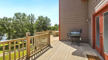 A deck with a barbecue grill, wooden railing, and a view of trees and a body of water.