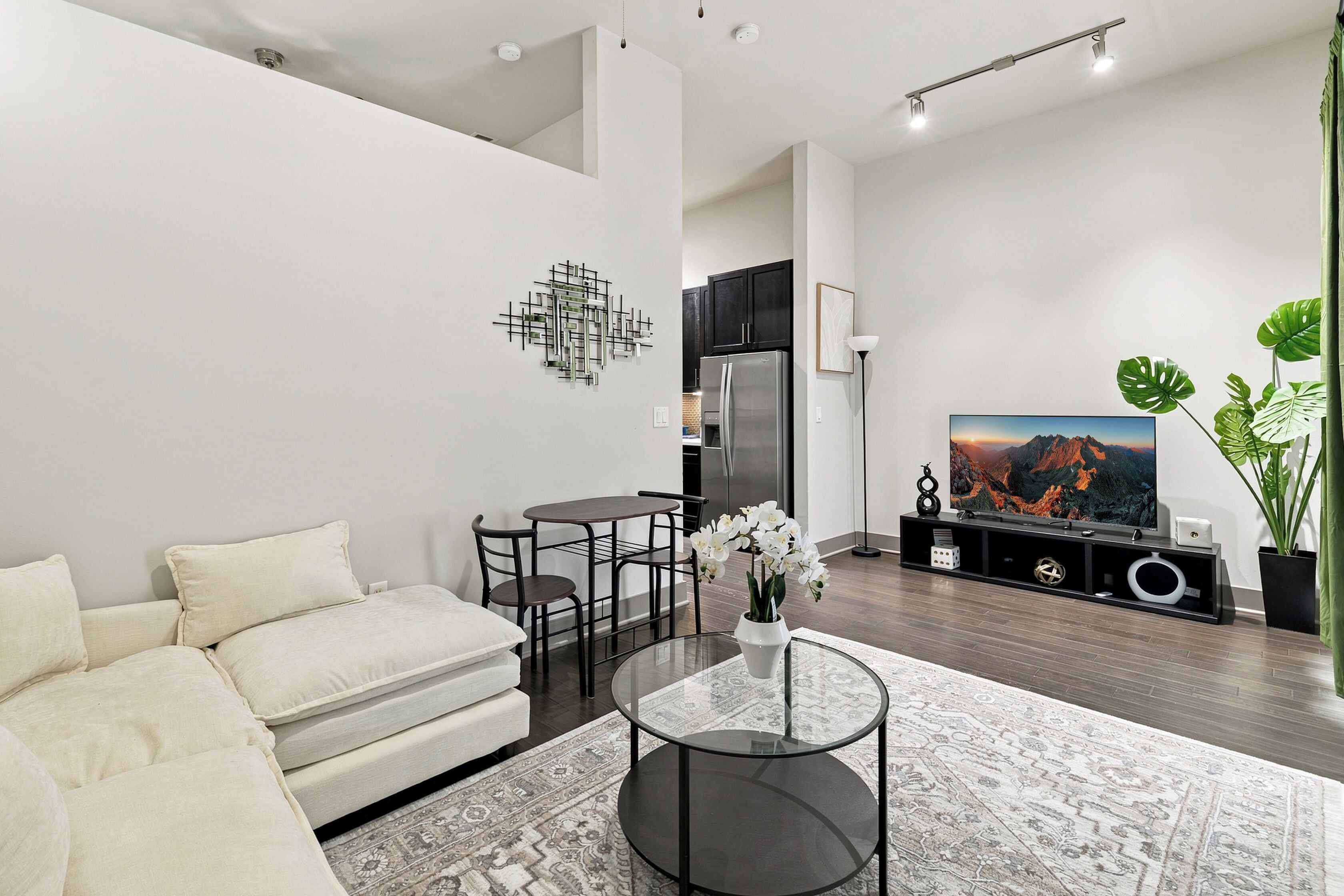 Ein modernes Wohnzimmer mit einer Couch, einem Couchtisch, einem Essbereich mit Stühlen, einer Küche mit Schränken, einem Kühlschrank, einem Flachbildfernseher und dekorativen Pflanzen.