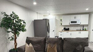 A modern living room with a grey sofa, a potted plant, and a kitchen in the background. - Modern 2BR Retreat | Cozy, Clean & Family-Friendly (Steinbach)