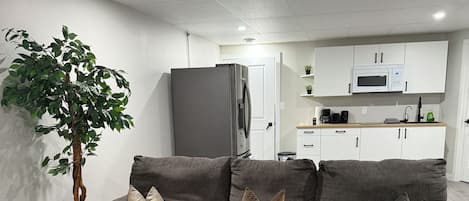 A modern living room with a grey sofa, a potted plant, and a kitchen in the background.