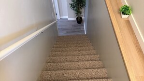 A carpeted staircase with a handrail, a potted plant, and a shelf with small plants. - Modern 2BR Retreat | Cozy, Clean & Family-Friendly (Steinbach)