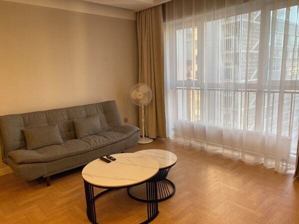 A living room with a sofa, a round table, a standing fan, and a large window with sheer curtains.