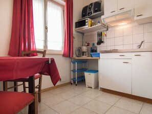 A compact kitchen with a dining area, including a table with a red tablecloth, a chair, and a window with red curtains. - Apartment 354 by Interhome (Fažana)