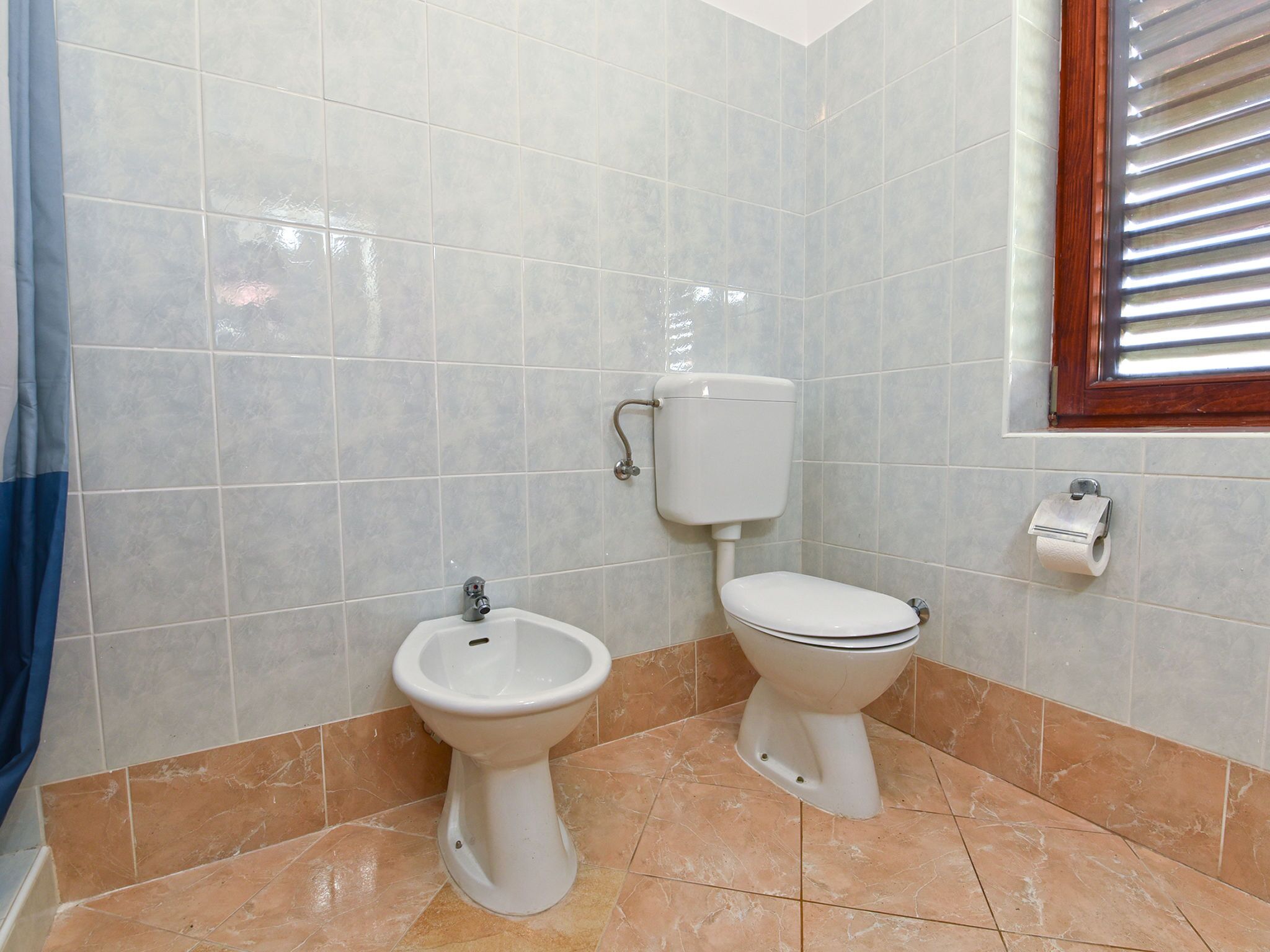 Un baño con bidé, inodoro y ventana con persianas.