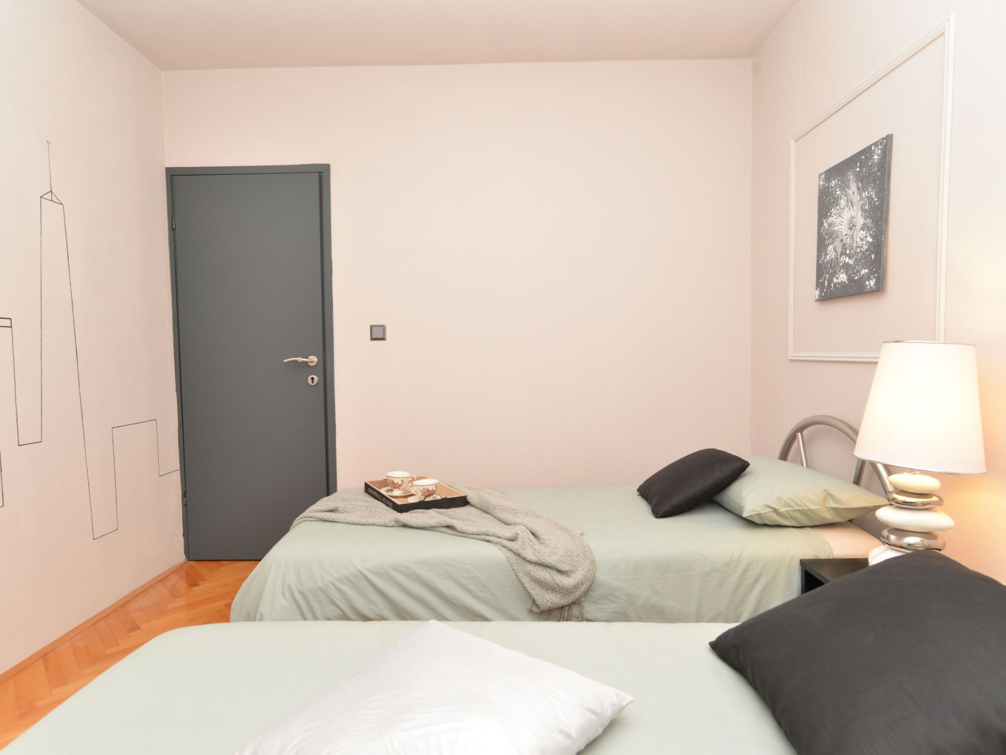 A bedroom with two beds, a door, a bedside table with a lamp, and a wall-mounted picture.