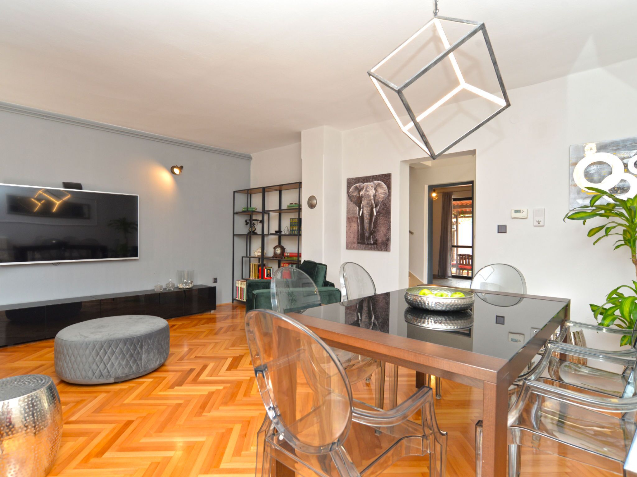 A modern living room with a flat-screen TV, a dining table with chairs, a glass coffee table, and a decorative elephant painting.