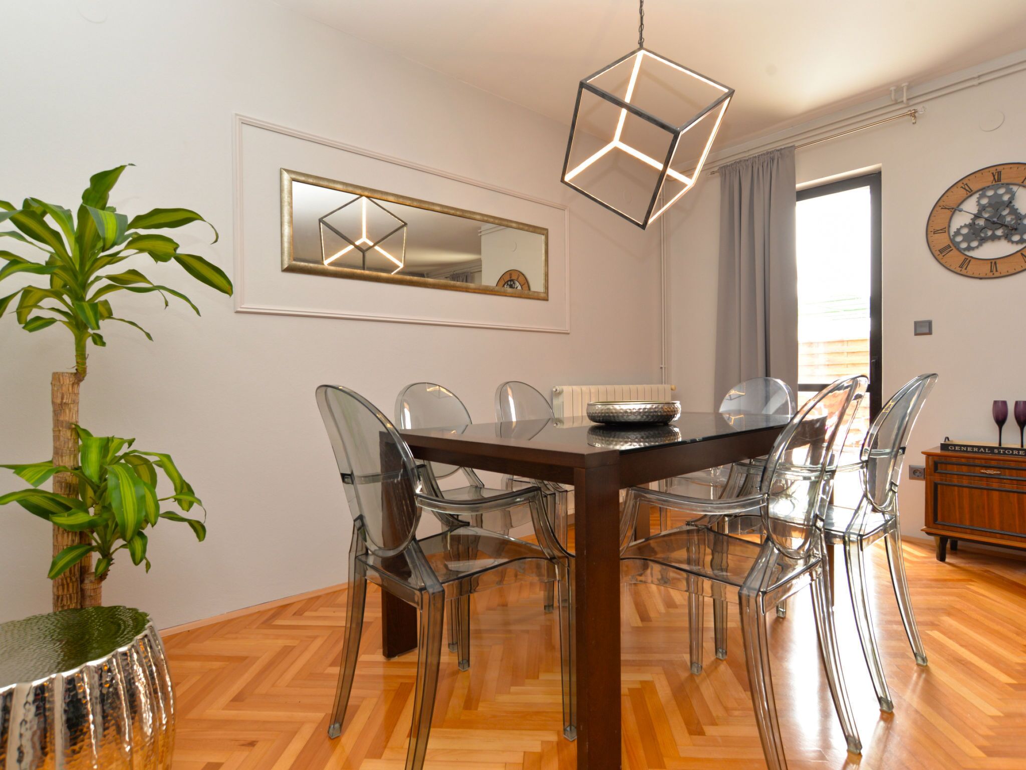 A dining area with a wooden table, clear chairs, a hanging light fixture, a mirror, and a clock on the wall.