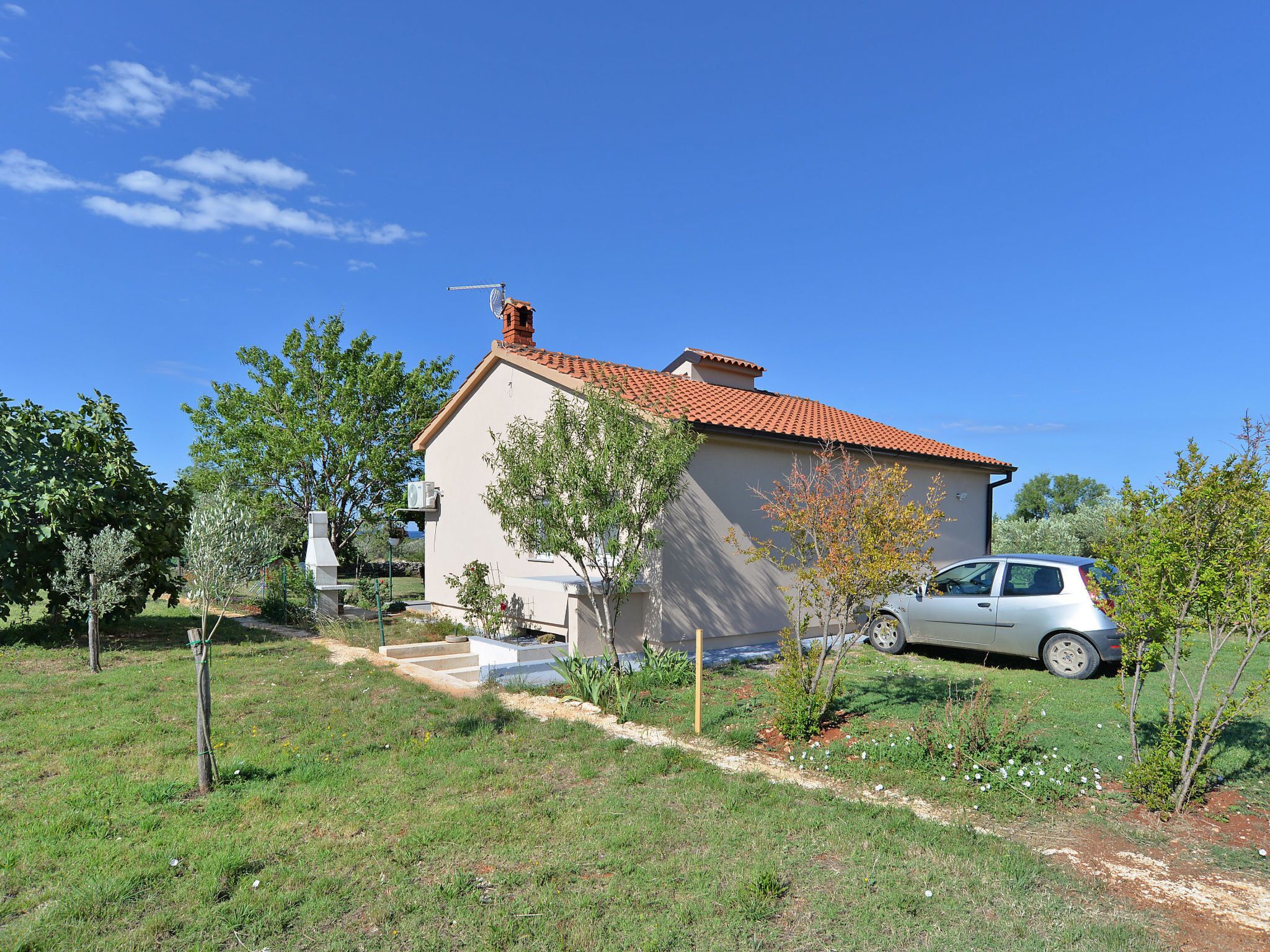 A single-story house with a red-tiled roof, a small garden with various plants, and a parked car in front.