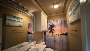 A bathroom with a bathtub, toilet, sink, and a shower curtain depicting a beach scene. - Welcome to Magnolia Pearls, a peaceful retreat for comfort and relaxation. (Prairieville)