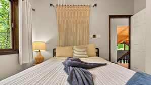 Un dormitorio con cama, ventana con cortinas, lámpara, puerta y vista a follaje exterior.