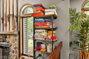 Een kamer met een stenen open haard, een houten bank, een boekenplank vol spelletjes en een groot raam met jaloezieën.