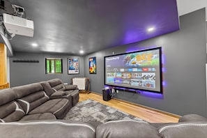 A modern home theater room with a large flat-screen TV, a leather recliner, and a projector mounted on the ceiling. - Poconos Chalet: Hot Tub, Theater, EV & Lake Access (Pocono Lake)