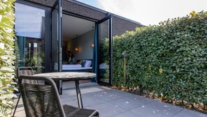 Eine offene Terrasse mit Tisch und Stuhl, ein Bett ist durch eine Glastür sichtbar, und im Hintergrund ein Heckenstreifen.