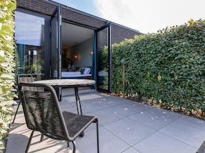 An open patio with a table and chair, a bed visible through a glass door, and a hedge in the background. - Studio RustUit by Interhome (Koudekerke)