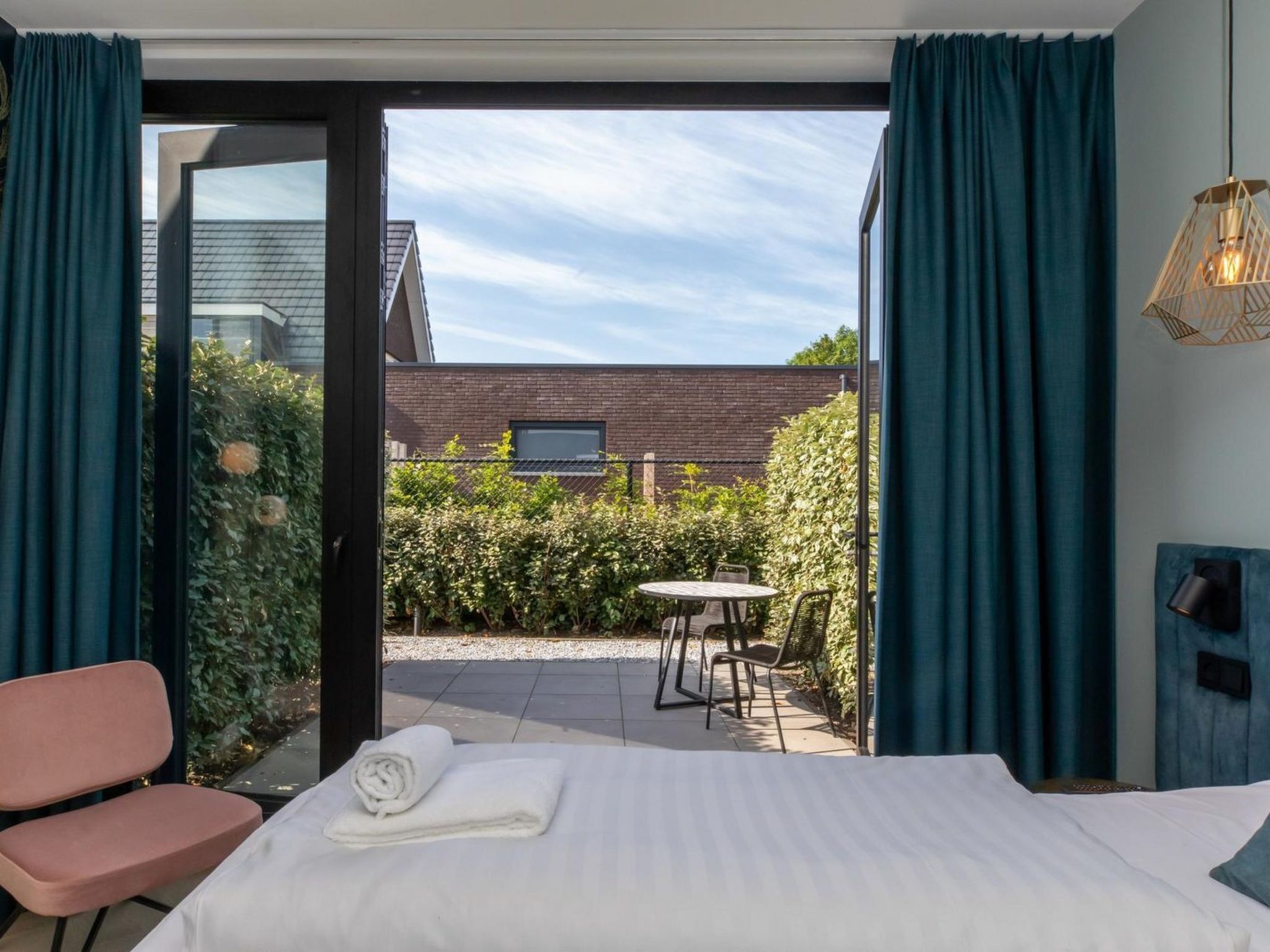 A bedroom with a bed, a chair, a small table, and a view of a garden and building outside.