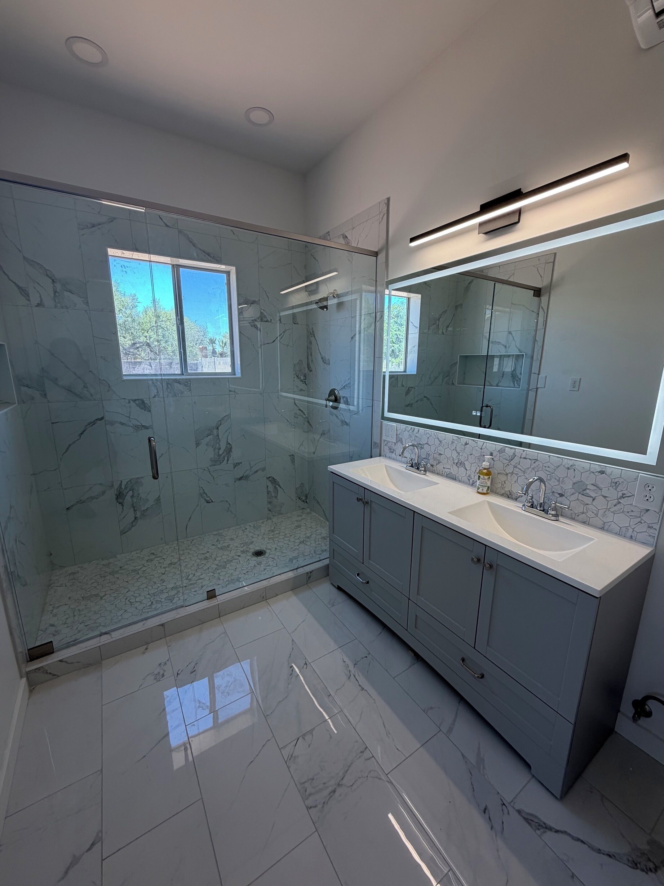 Un baño moderno con ducha envuelta en vidrio, un mueble de tocador amplio con lavamanos doble y un espejo grande sobre el mueble.