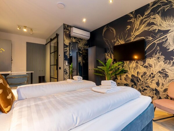 Habitación de hotel con dos camas, un televisor de pantalla plana montado en la pared, un equipo de aire acondicionado y una planta grande en maceta.