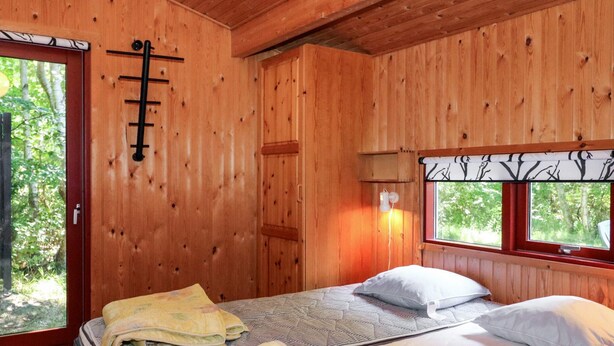 A wooden cabin room with a bed, two pillows, a window with a tree view, and a door with a ladder. - 8 Person Holiday Home in Hadsund (Hadsund)