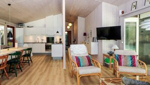 A modern kitchen with white cabinets, a dining area with wooden chairs, a sofa with colorful cushions, and a TV mounted on the wall. - 8 Person Holiday Home in Hadsund (Hadsund)
