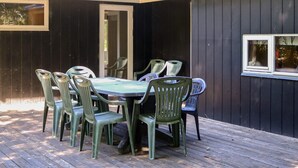 An outdoor patio with a table and chairs, a wooden deck, and a dark-colored building with a window. - 8 Person Holiday Home in Hadsund (Hadsund)