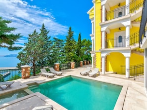 Ein Poolbereich mit Liegestühlen, freier Blick auf das Meer und ein gelbes Gebäude mit einem Balkon.
