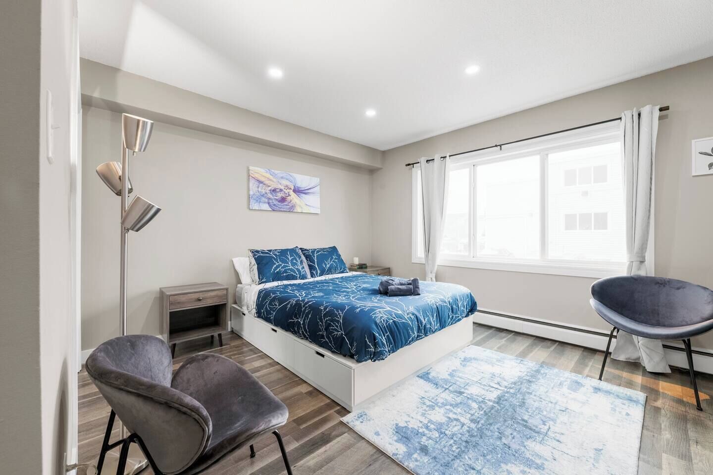A modern bedroom with a bed, bedside tables, a chair, a floor lamp, and a large window.