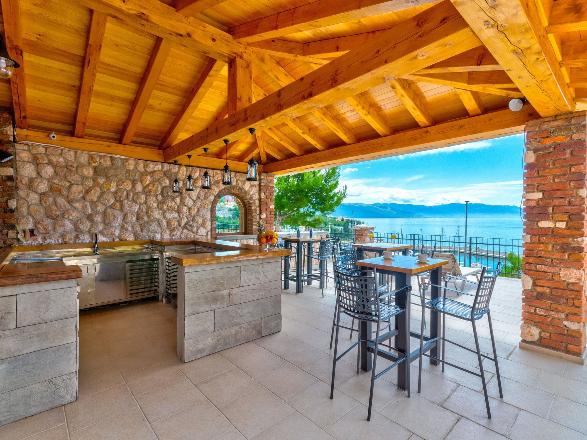An outdoor bar area with stone walls, wooden ceiling beams, and a view of the sea.