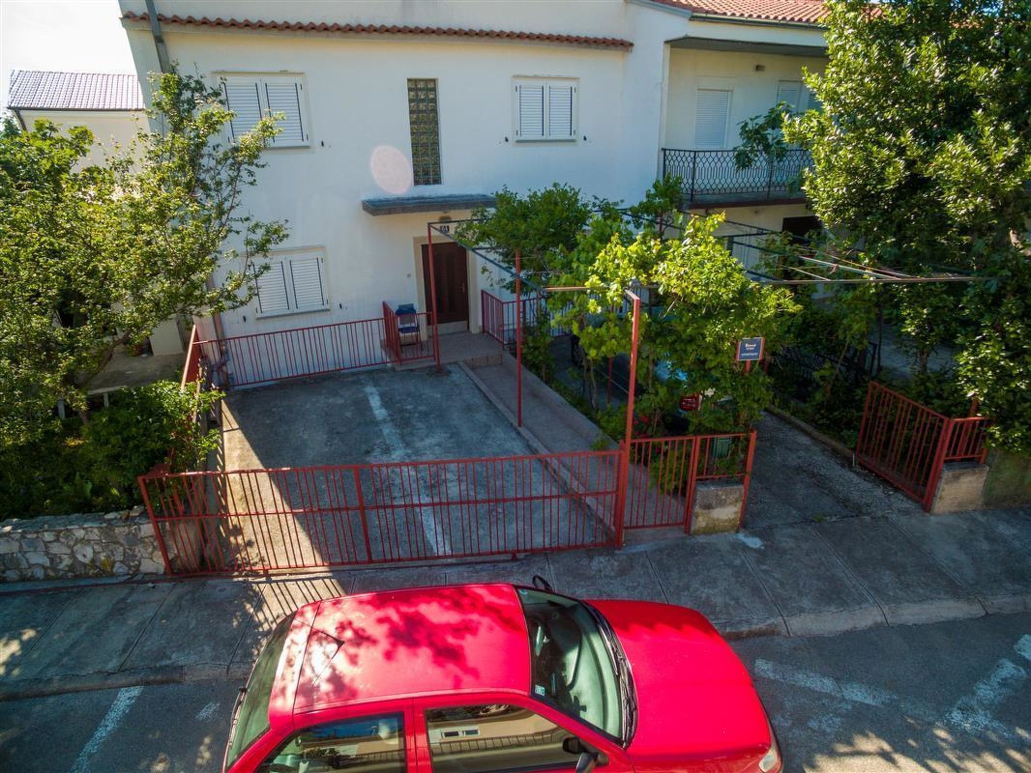 A red car parked in front of a building with a gated entrance, a staircase leading to a balcony, and a tree in the foreground.