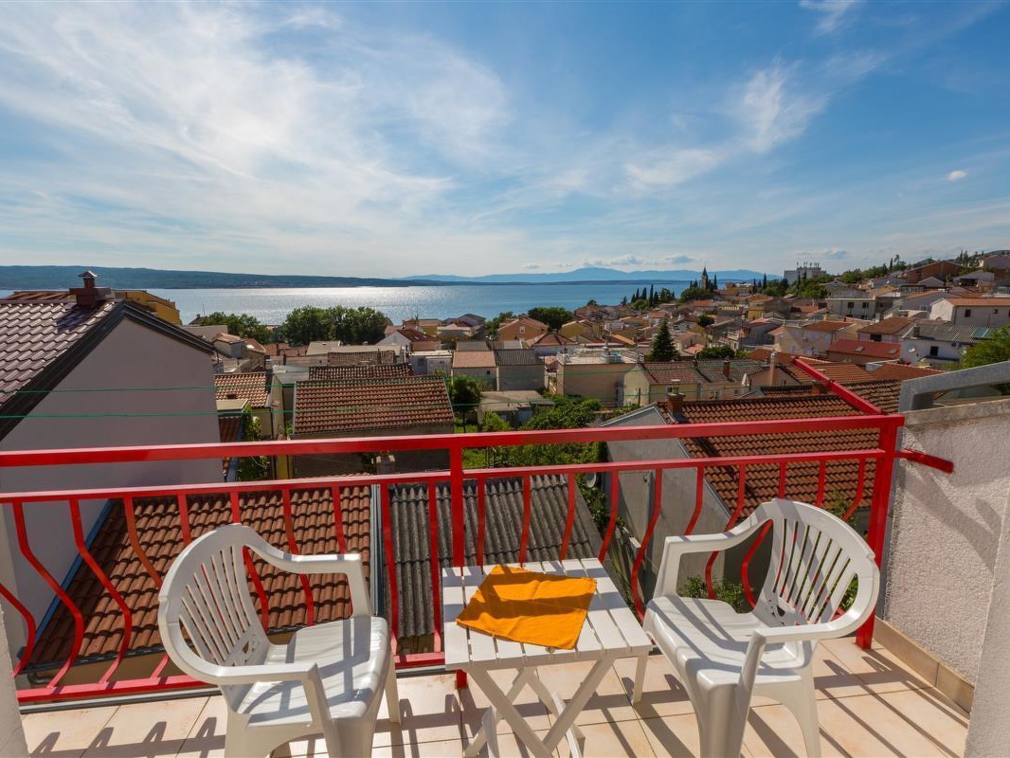 A balcony with two white chairs and a small table, overlooking a town with a body of water and mountains in the distance.