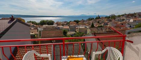 A balcony with two white chairs and a small table, overlooking a town with a body of water and mountains in the distance.