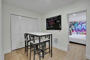 A room with a white table and chairs, a closed white closet, a wall with a blackboard, and a door leading to a room with graffiti art. - MiamiMuralHouse:4BRw/Themes, Gameroom, Backyard (Miami)
