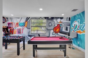 A pool table with a red felt top, a foosball table, and a wall with sports-themed murals including a basketball and a Miami Heat logo. - MiamiMuralHouse:4BRw/Themes, Gameroom, Backyard (Miami)