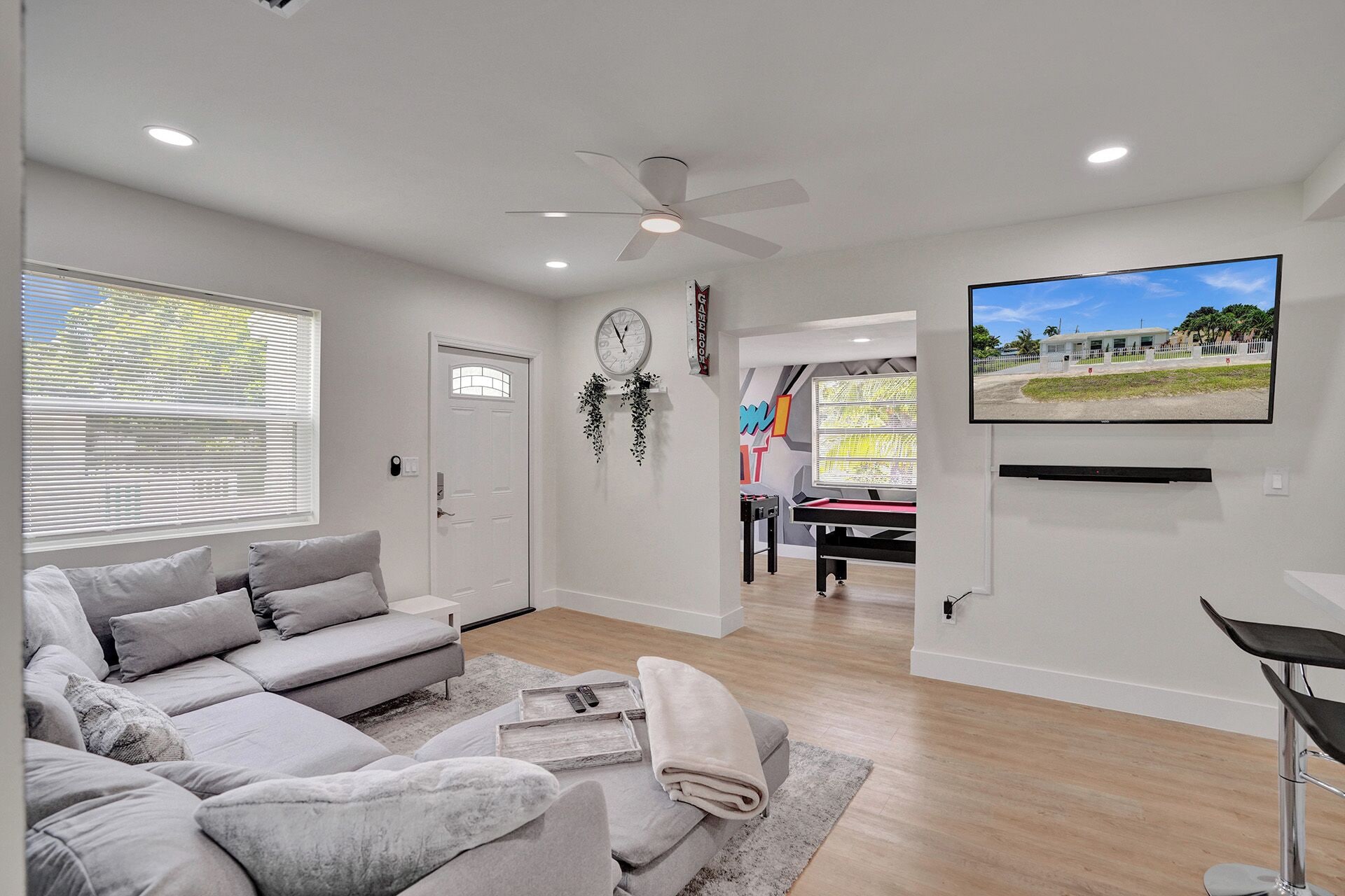 A living room with a grey sofa, a ceiling fan, a TV mounted on the wall, and a door with a window.