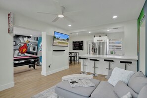 A modern living room with a grey sofa, a TV, and a kitchen area with a fridge and bar stools. - MiamiMuralHouse:4BRw/Themes, Gameroom, Backyard (Miami)