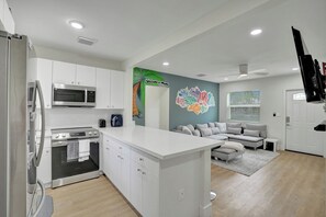 A modern kitchen with white cabinetry, a built-in microwave, and an oven. There is a dining area with a gray sofa and a small table. A colorful wall mural is visible, and a ceiling fan is installed. - MiamiMuralHouse:4BRw/Themes, Gameroom, Backyard (Miami)