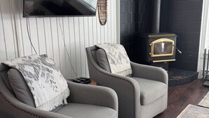 Ein Wohnzimmer mit zwei grauen Sesseln, einem Flachbildfernseher, einem schwarzen Kamin und einem gemusterten Teppich.