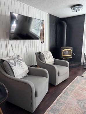 Ein Wohnzimmer mit zwei grauen Sesseln, einem Flachbildfernseher, einem schwarzen Kamin und einem gemusterten Teppich.