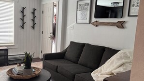 Ein Wohnzimmer mit einem dunkelgrauen Sofa, einem runden Couchtisch mit einem gewebten Tablett, einem gemusterten Teppich und einer weißen Tür.